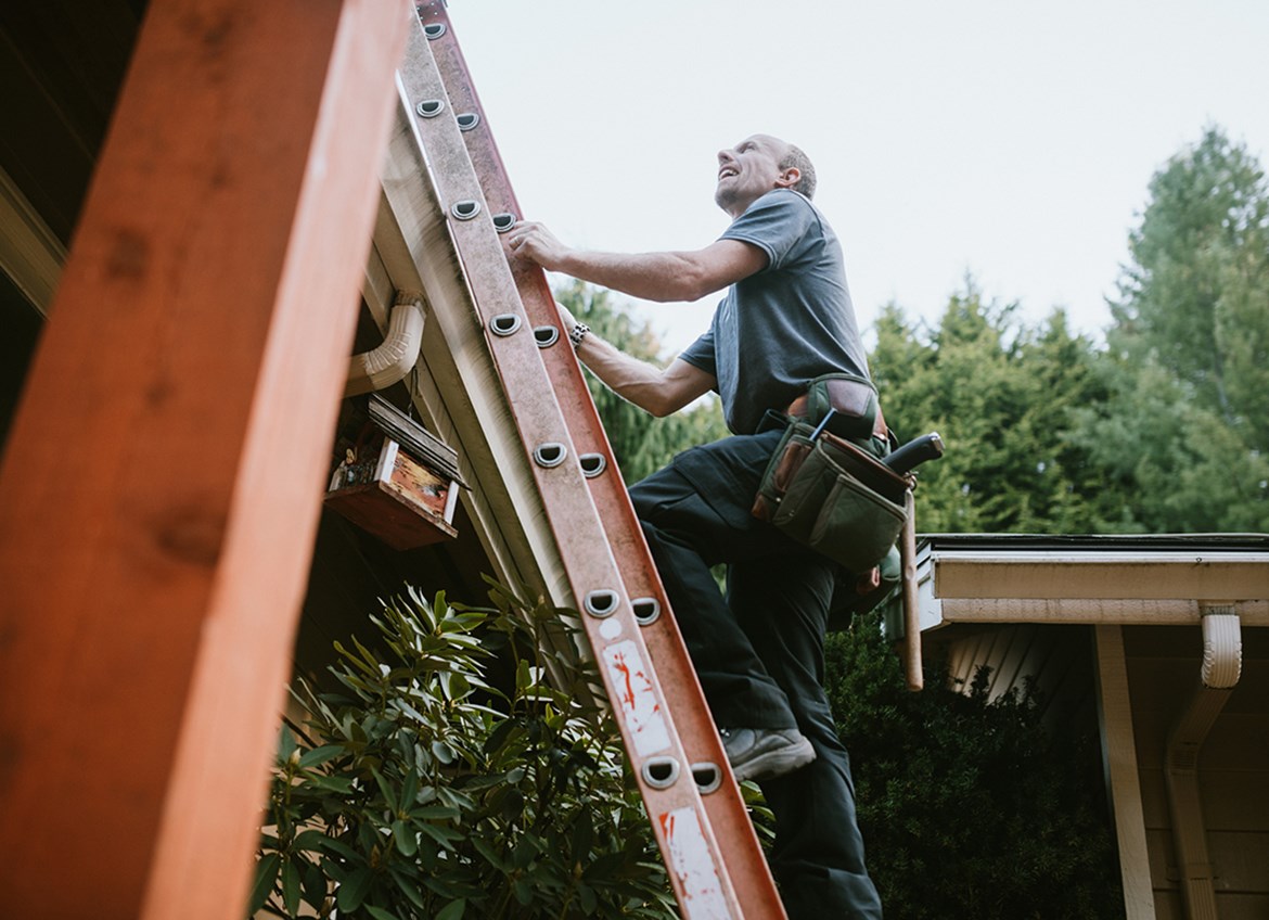 Man wearing tool belt climbs ladder onto roof