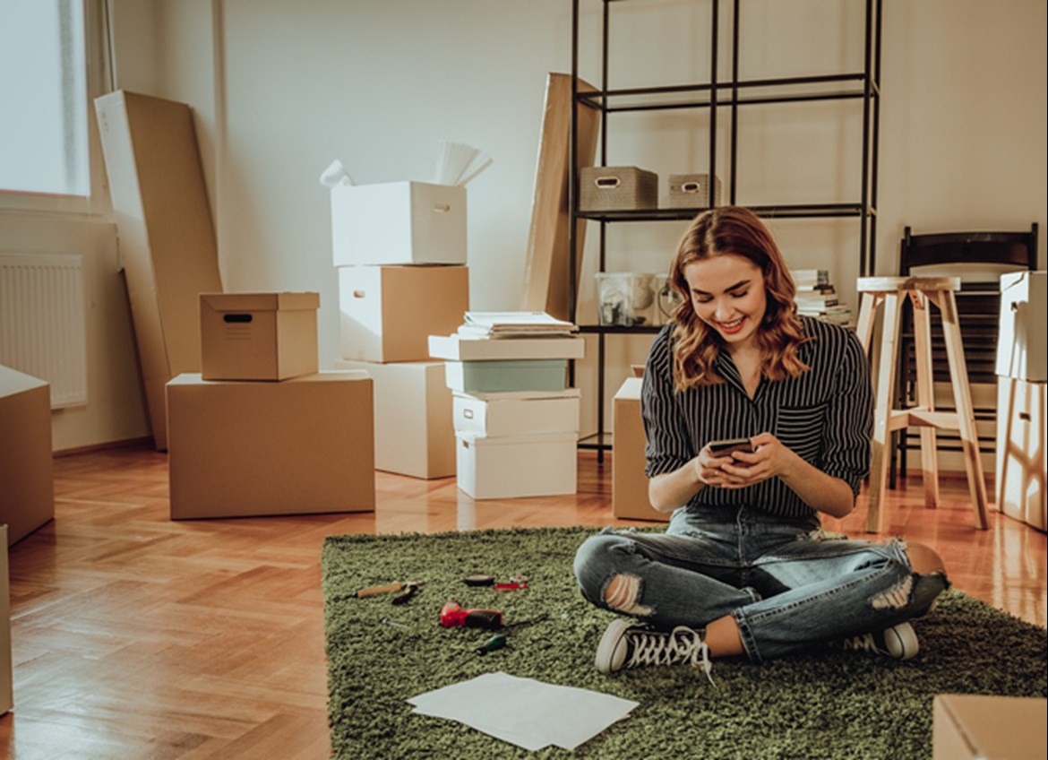 Woman sitting on floor looking at phone with boxes surrounding her