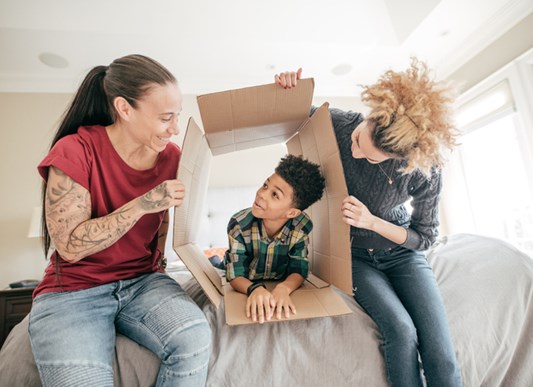 Two women holding out box on bed with little boy playing inside